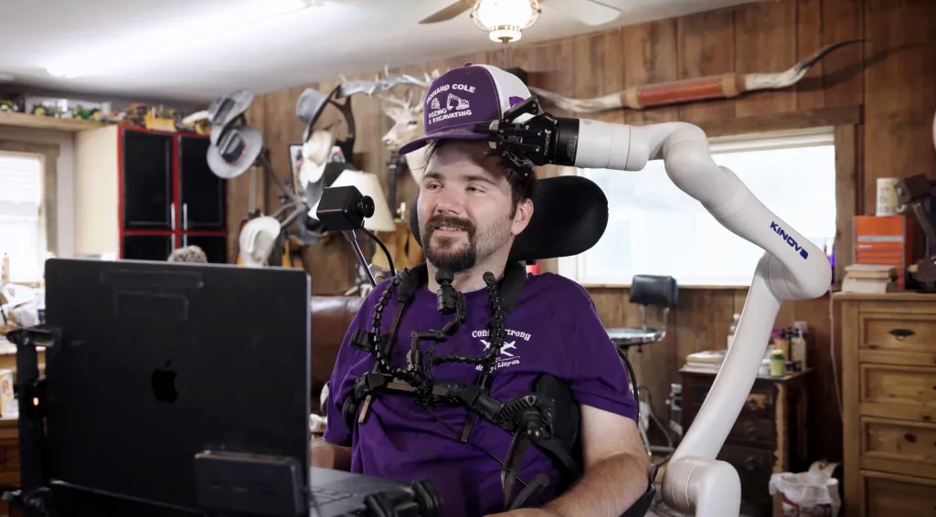 A man wearing a purple hat and shirt, with a robotic arm attached to his hat, looks toward a laptop.