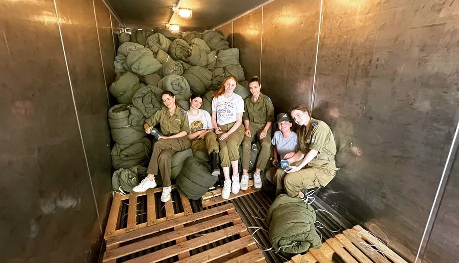 Six young women, some in military uniforms, sit on wooden pallets and green rolled sleeping bags inside a metal storage unit.