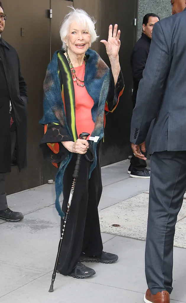 Ellen Burstyn waves, holding a cane, wearing a bright orange top and a multi-colored shawl.