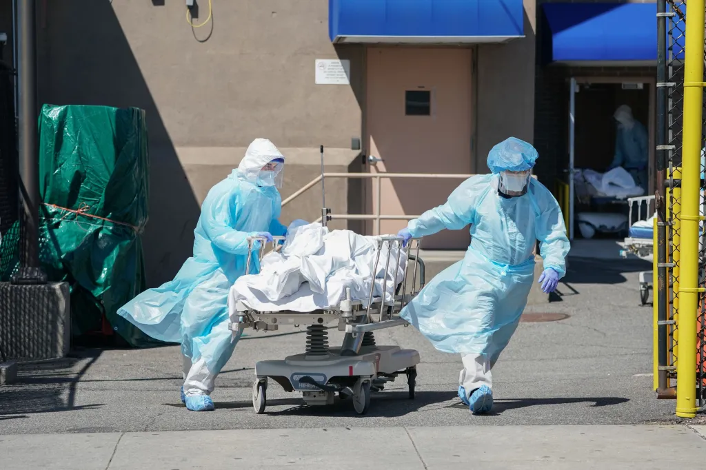 Healthcare workers in PPE move a body on a gurney towards a temporary morgue.