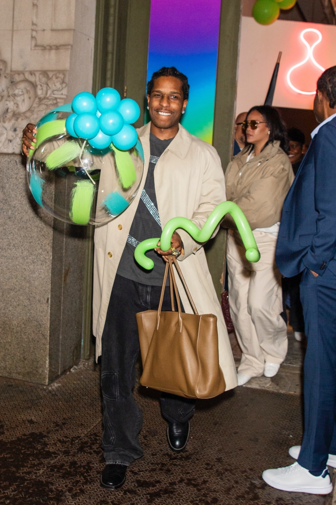 A$AP Rocky smiling and holding a clear balloon with blue and green accents and a green balloon animal, with Rihanna visible in the background.