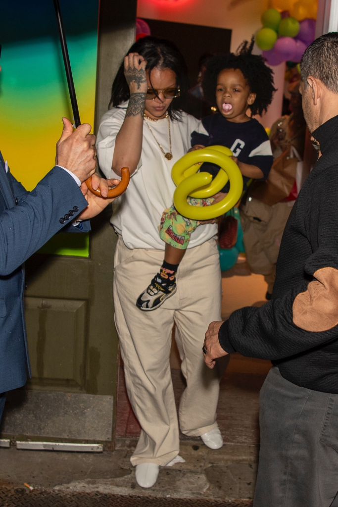 Rihanna carrying RZA, who is holding a yellow balloon animal, while a person holds an umbrella and another person stands to their right.