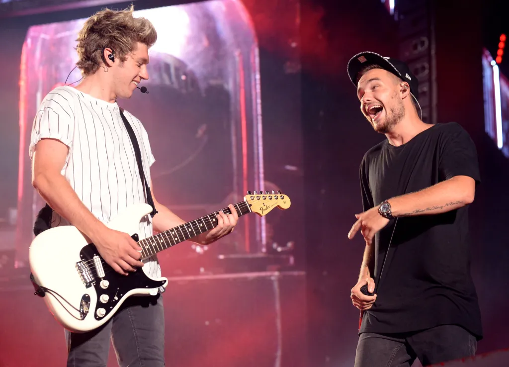 Niall Horan (left) smiles while playing guitar and Liam Payne (right) laughs while holding a microphone and pointing at Horan during a performance.