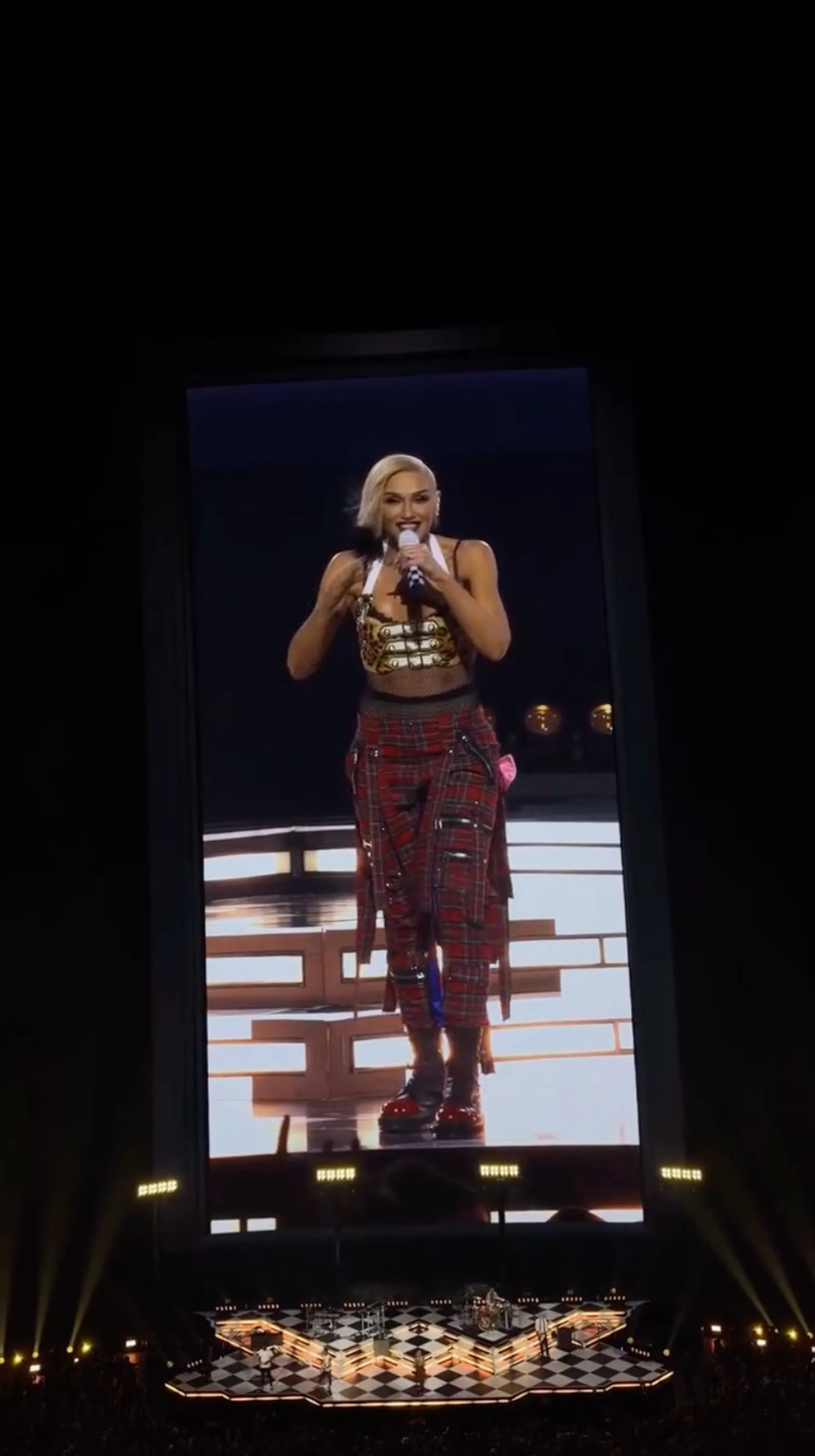 Gwen Stefani performing on stage, wearing a gold and white top with red plaid pants and boots.