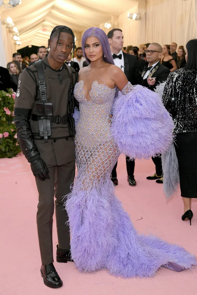 Travis Scott in brown military-inspired outfit and Kylie Jenner in a lavender feathered and embellished dress on the pink carpet of the 2019 Met Gala.