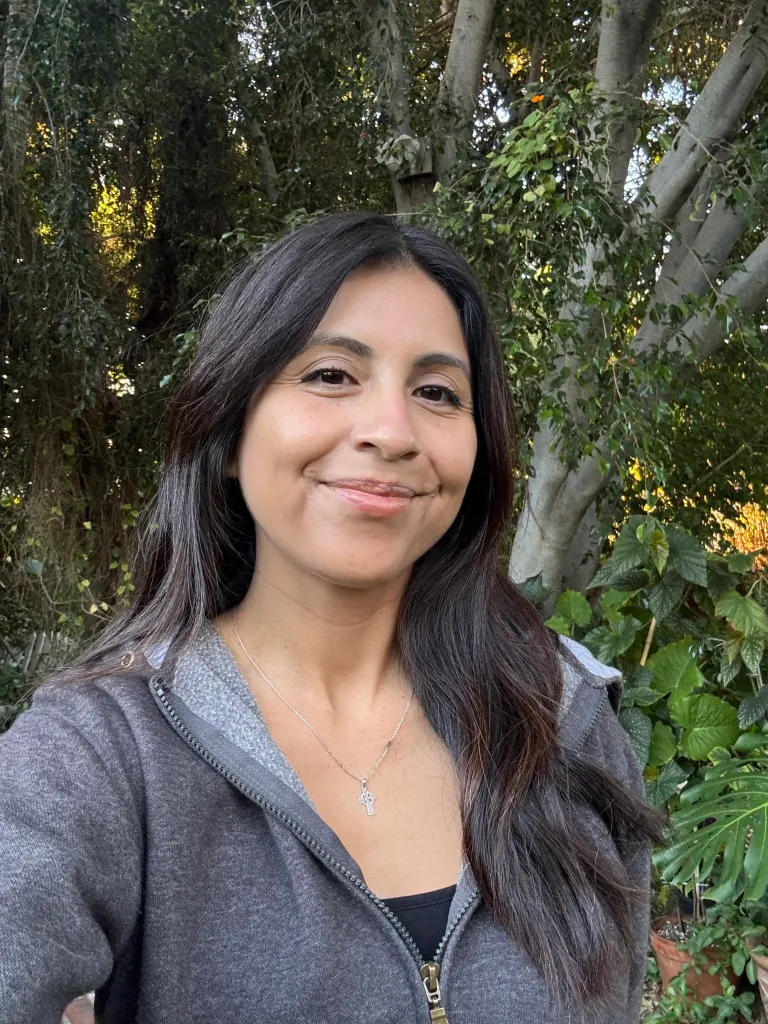 Shaina Montiel, a hantavirus survivor, smiles at the camera in an outdoor setting with trees and foliage in the background.
