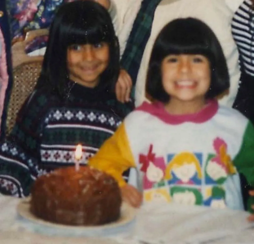 Shaina (L) as a child, with another girl, smiling behind a birthday cake.