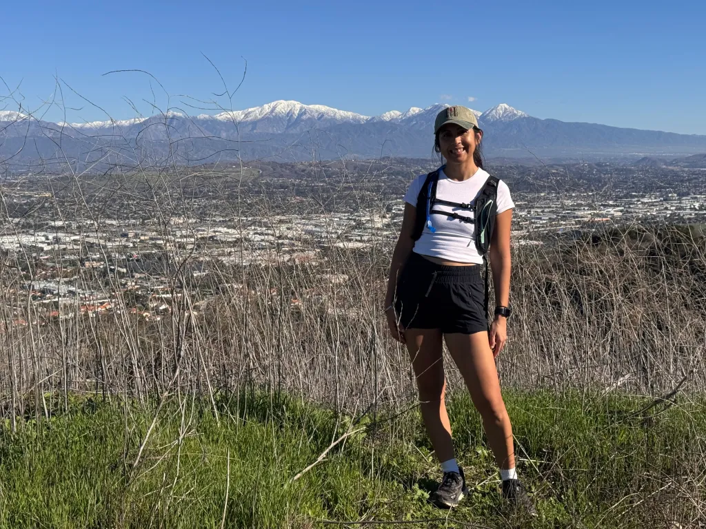 A woman smiling while standing on a grassy hill overlooking a city with snow-capped mountains in the background.