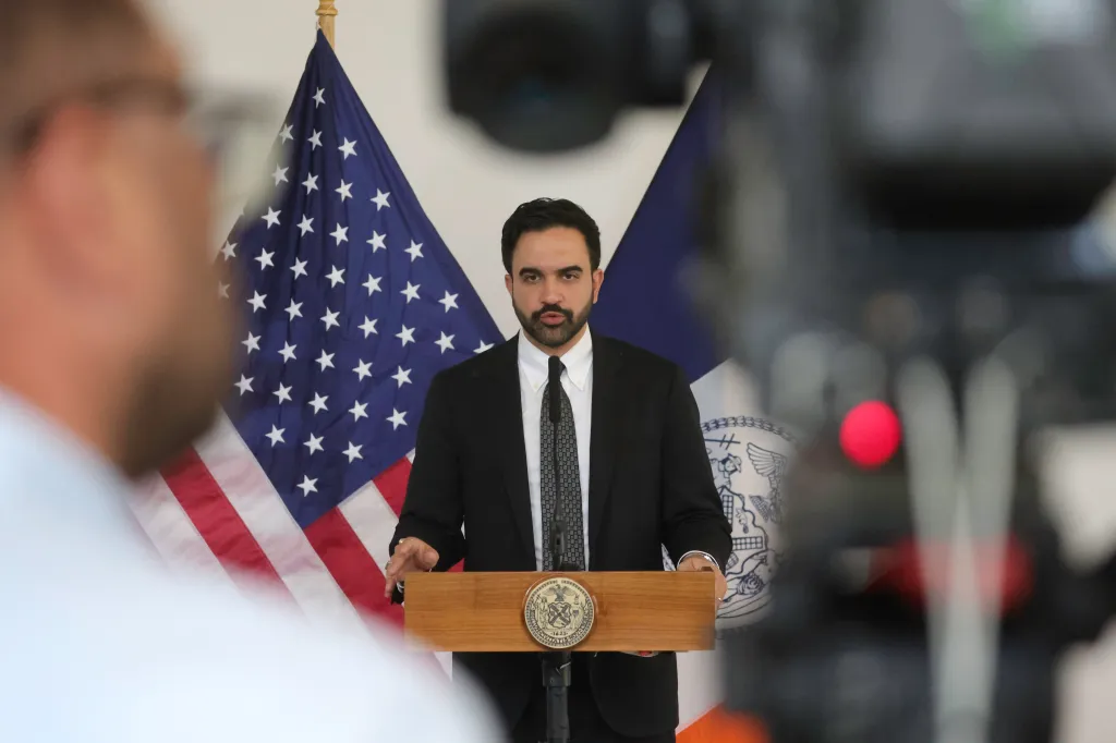 NYC Mayor Zohran Mamdani speaking at a podium with American and New York City flags behind him.