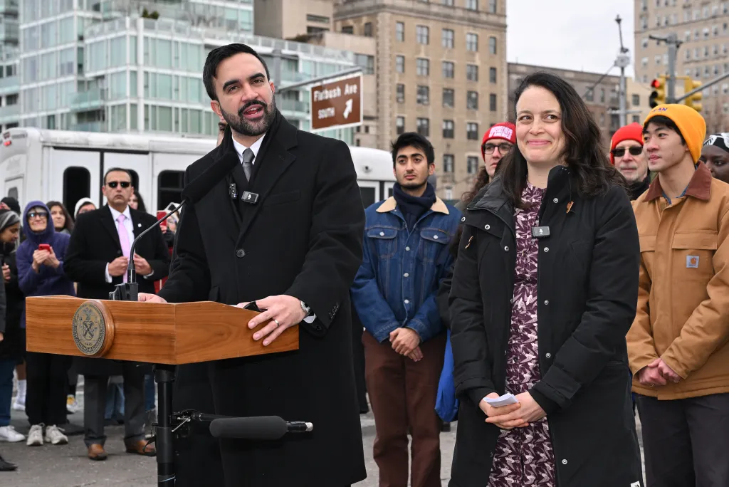 NYC Mayor Zorhan Mamdani speaking at a podium with Tascha Van Auken standing next to him.