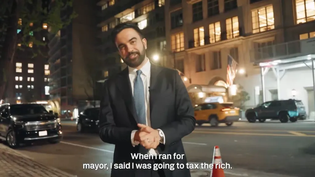 Zohran Mamdani, in a suit, stands on a street at night with city buildings and cars in the background, speaking about taxing the rich.