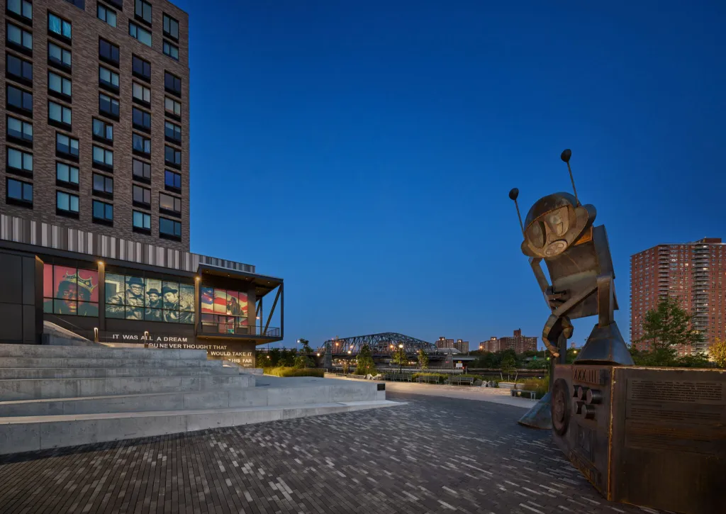 Exterior view of the Universal Hip Hop Museum in the Bronx, New York, featuring large-scale portraits of hip-hop artists, with a metallic statue of a boombox and character on the right.