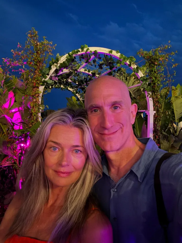 Paulina Porizkova and Jeff Greenstein smiling together at night, illuminated by pink and purple lights, with an arched trellis behind them.