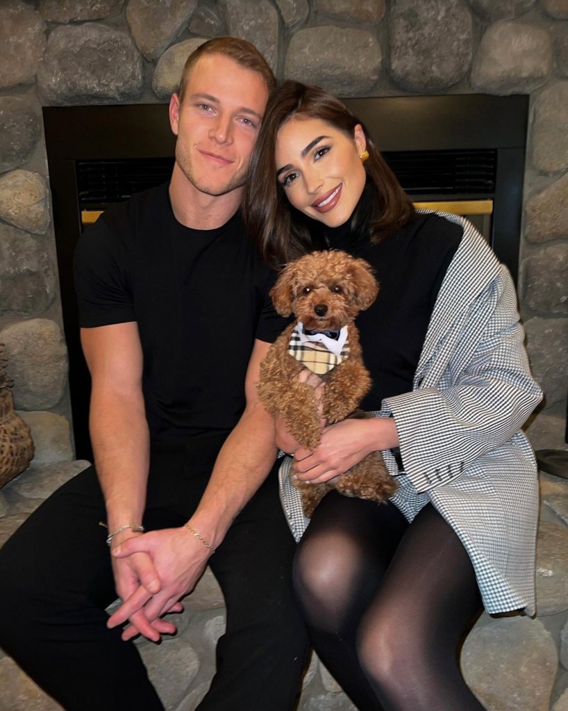 Olivia Culpo and Christian McCaffrey with a small dog in front of a stone fireplace.