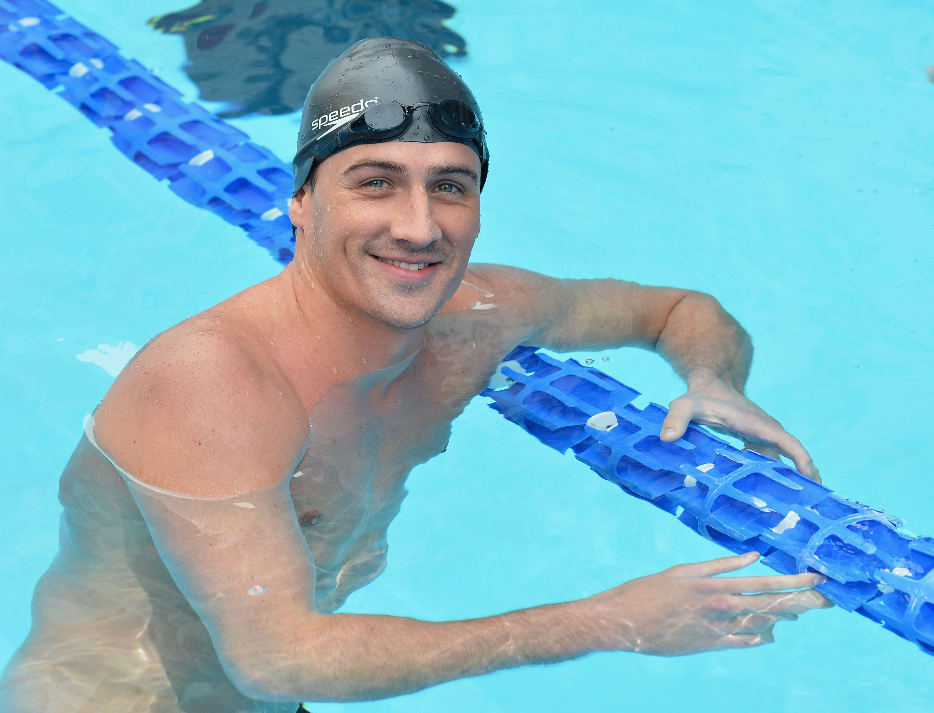 Olympic gold medalist Ryan Lochte smiling in a swimming pool, wearing a black swim cap and goggles, holding onto a blue lane line.
