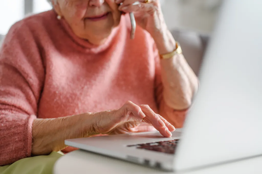 An elderly woman uses a laptop while talking on the phone.