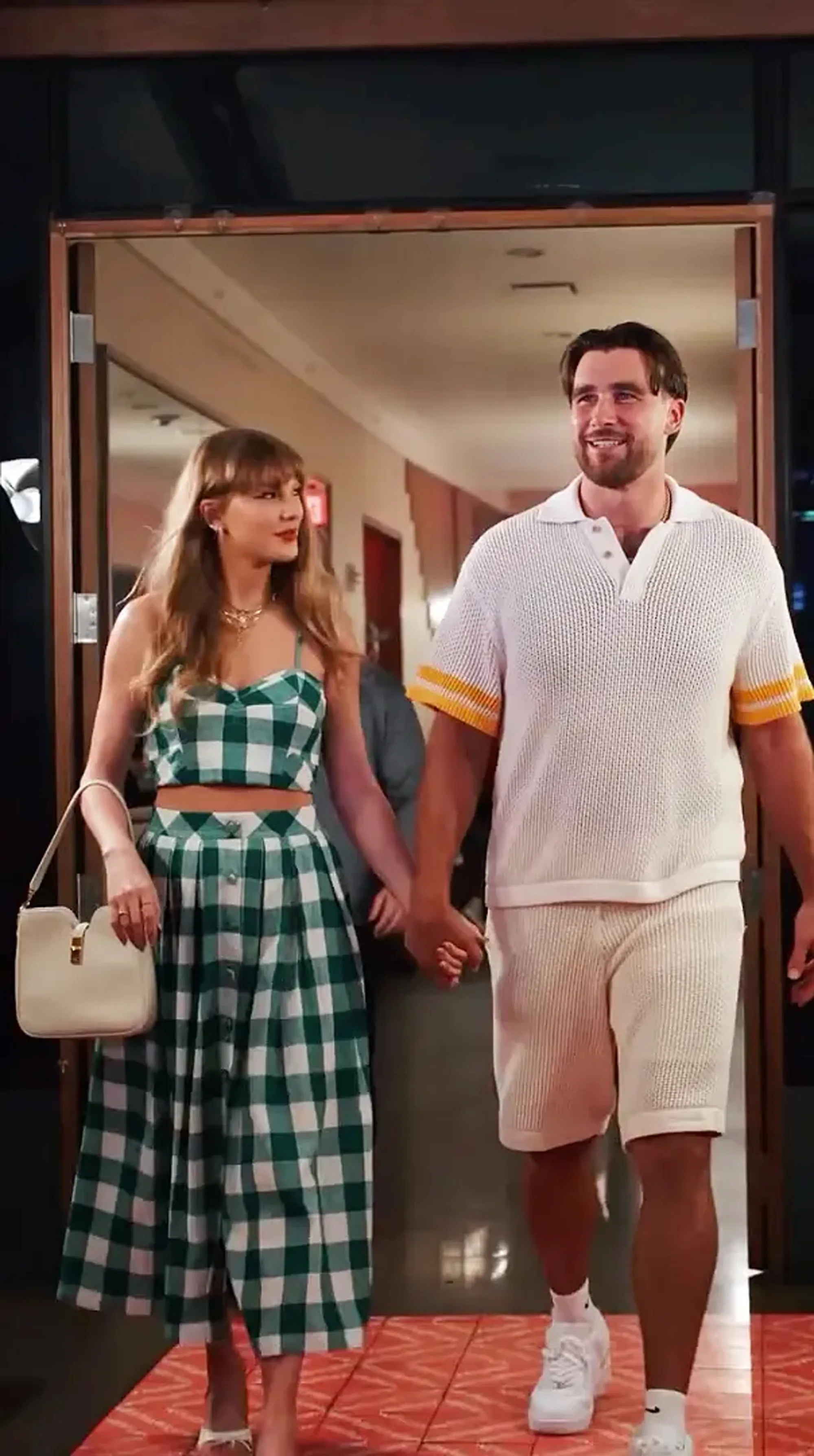 Taylor Swift in a green and white gingham outfit holding hands with Travis Kelce in a white polo and shorts.