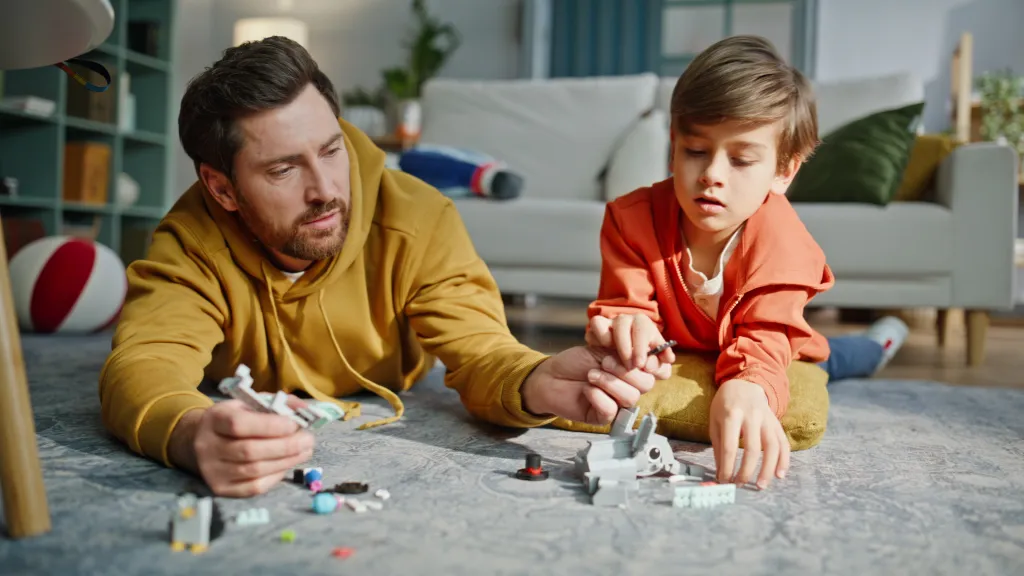 A father and son lying on a rug building a gray Lego creation together.