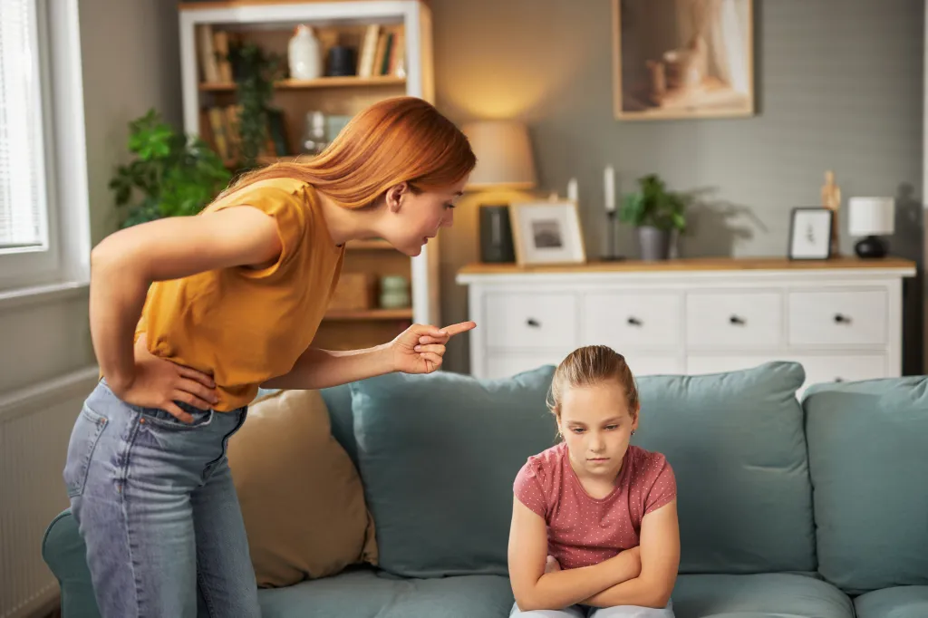 A parent scolding a child who is seated on a couch with her arms crossed.