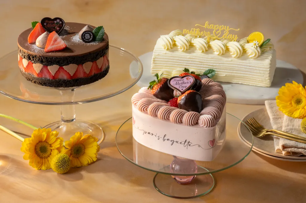 Three Paris Baguette Mother's Day special cakes, including chocolate with strawberries, a white layered cake, and a heart-shaped pink cake, displayed on stands with yellow flowers.