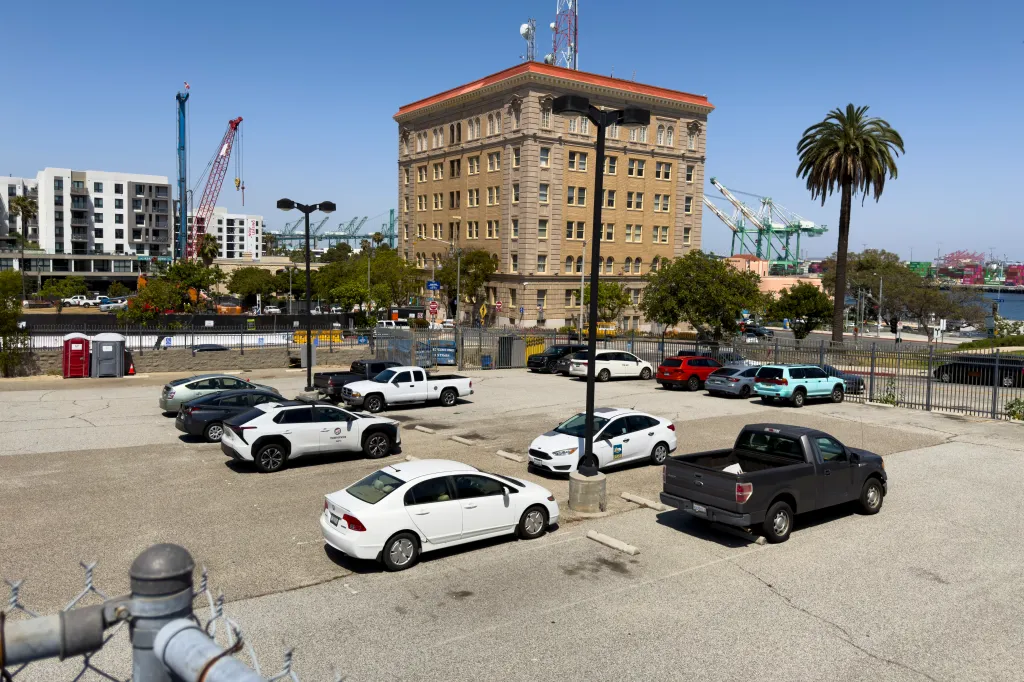 A parking lot at 711 S. Beacon St. in San Pedro, Los Angeles, showing an empty lot with several cars parked, a historic building, construction cranes, and the harbor in the background.