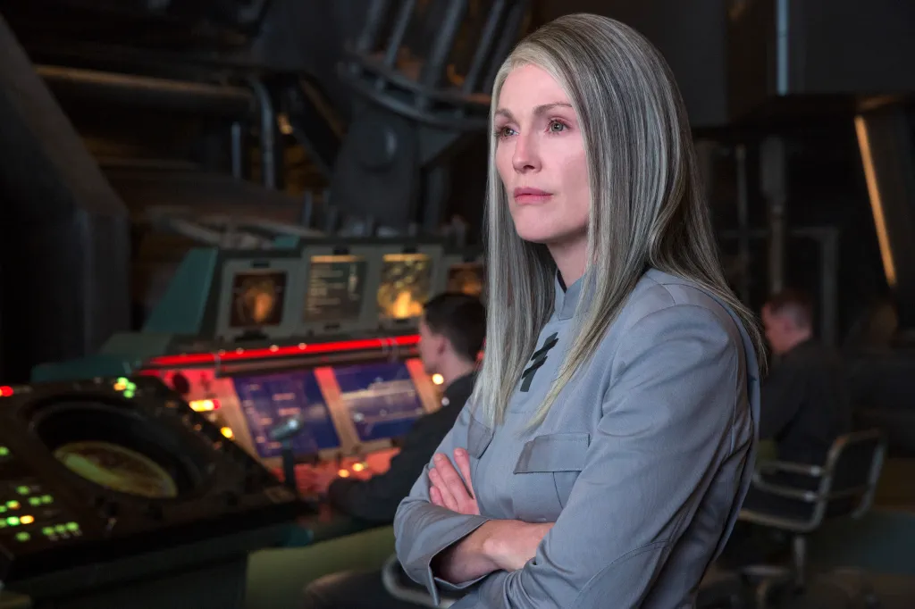 Julianne Moore as President Alma Coin with gray hair, wearing a gray uniform with a black insignia, standing with arms crossed in a control room.