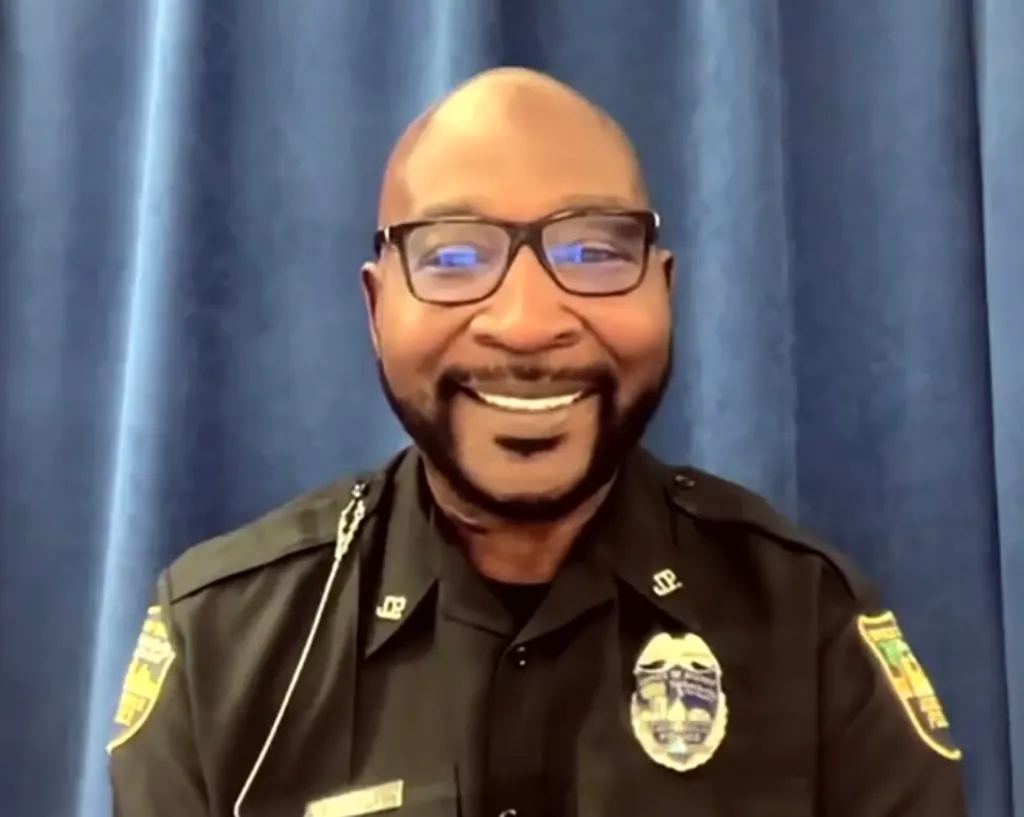 A bald, smiling male police officer wearing glasses and a uniform.