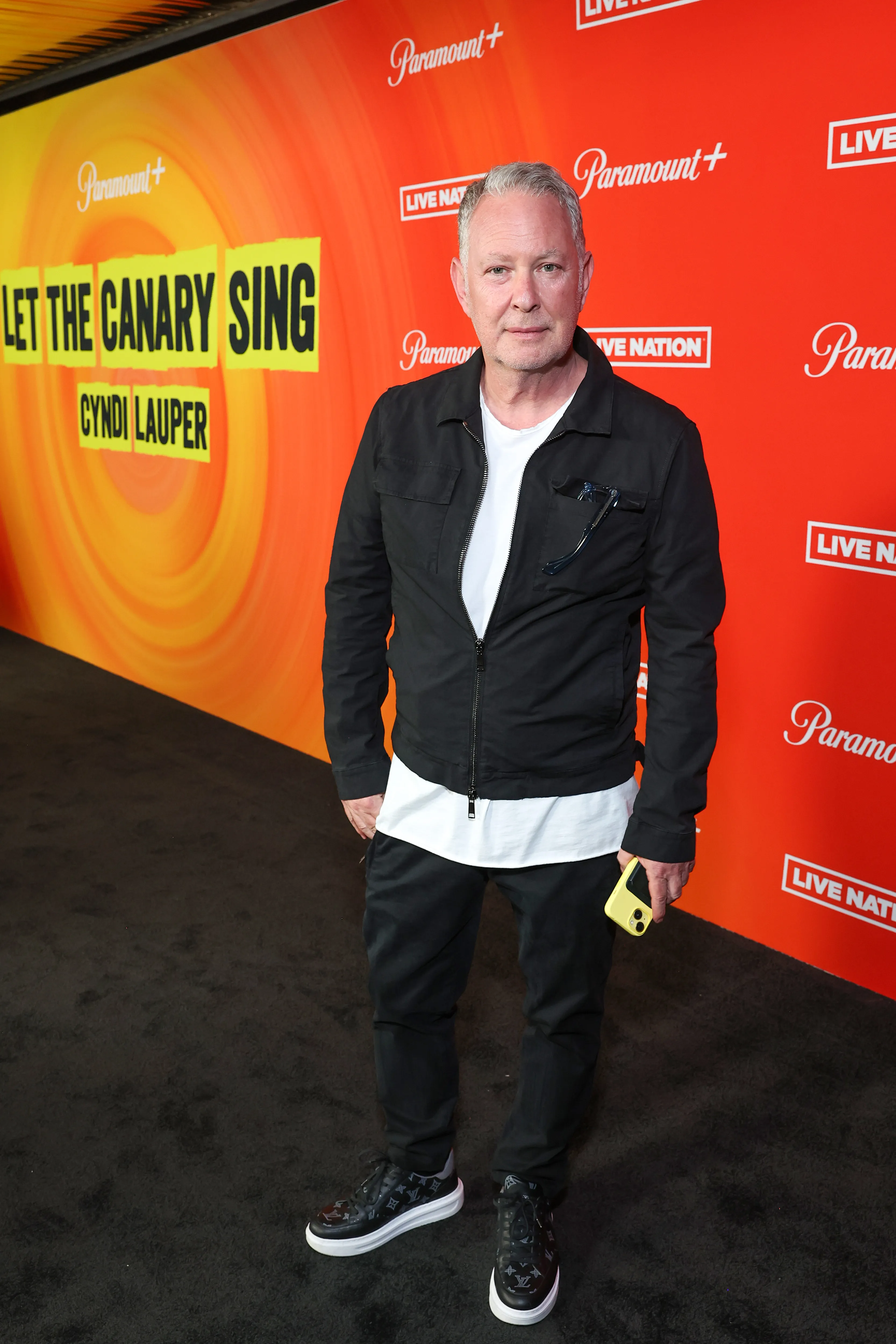Paul Kemsley standing on a black carpet in front of an orange and yellow background with 