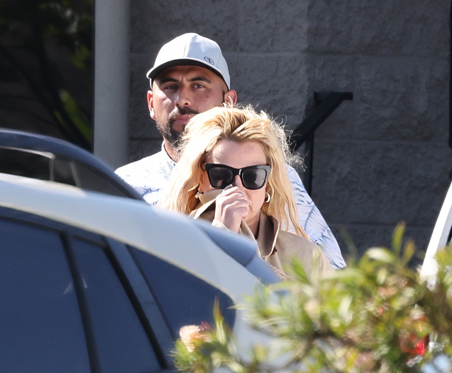 Britney Spears wearing sunglasses and a Burberry trench coat next to her driver, with a car and foliage in the foreground.