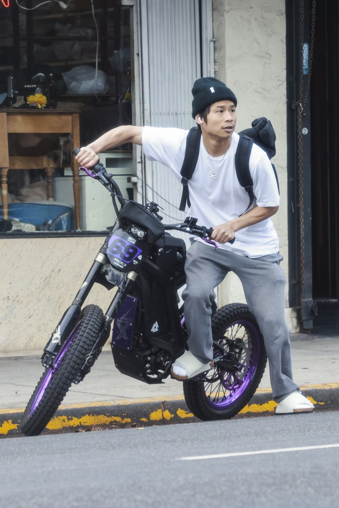 Pax Jolie-Pitt on a black and purple Talaria Sting R MX4 Electric Dirt Bike, wearing a white t-shirt, black hat, and black backpack, outside a Los Angeles smoke shop.