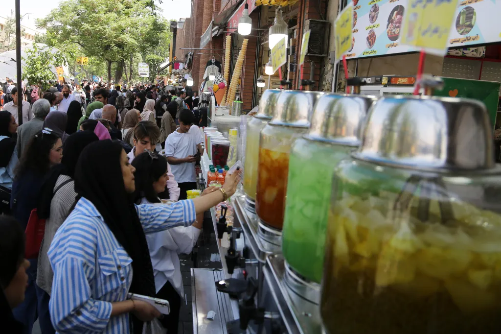 A woman in a blue and white striped shirt reaching for a drink from a street vendor in a crowded Tehran street.