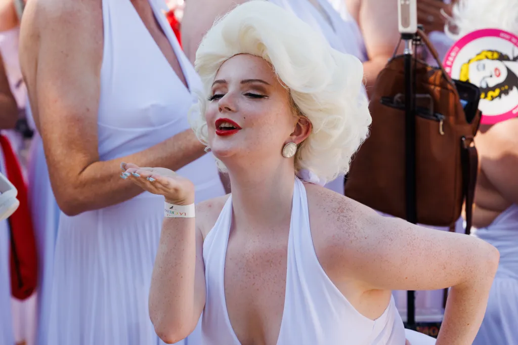 A person dressed as Marilyn Monroe, wearing a white halter dress and blonde wig, blowing a kiss to the camera.