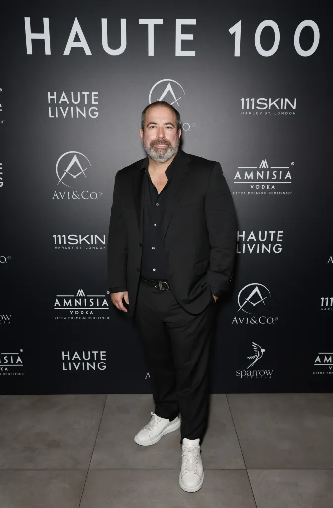 Manny Angelo Varas posing in a black suit with a white sneakers in front of a backdrop with various logos.