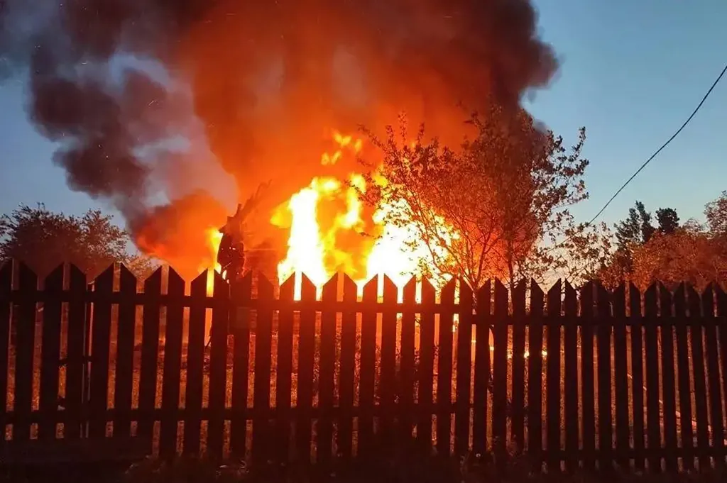 A building engulfed in flames and thick black smoke seen from behind a wooden fence.