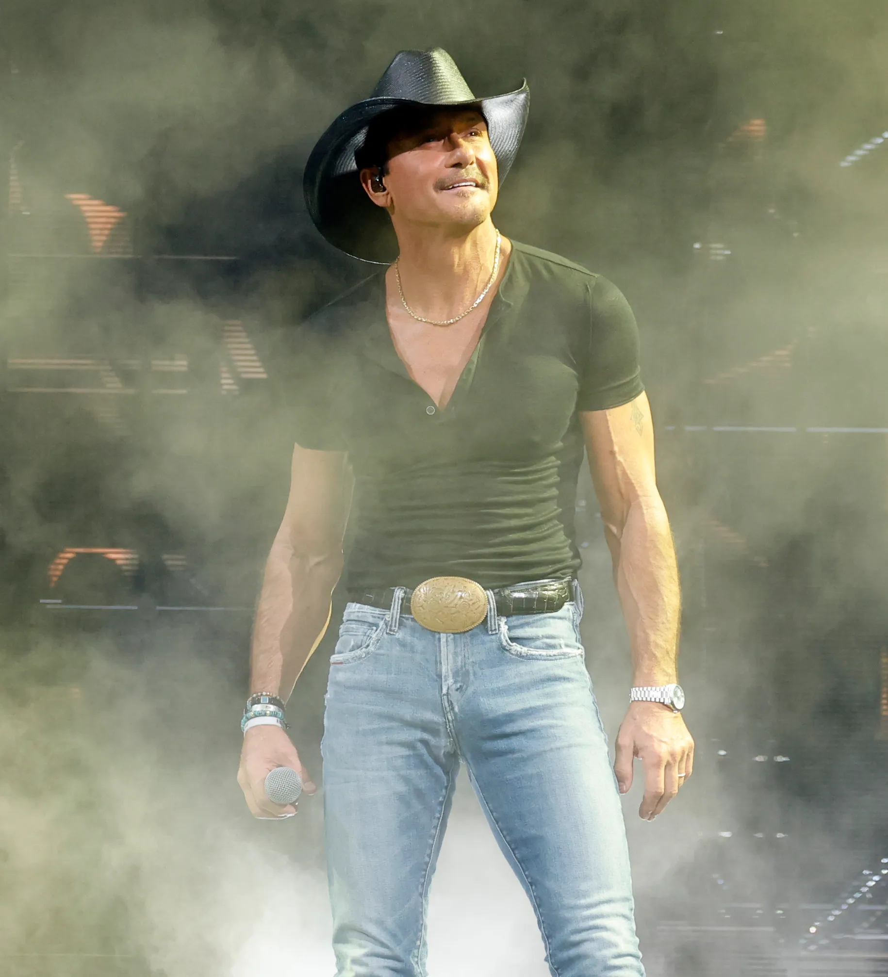 Tim McGraw performing on stage, wearing a black cowboy hat, black short-sleeved shirt, blue jeans, and a large gold belt buckle.