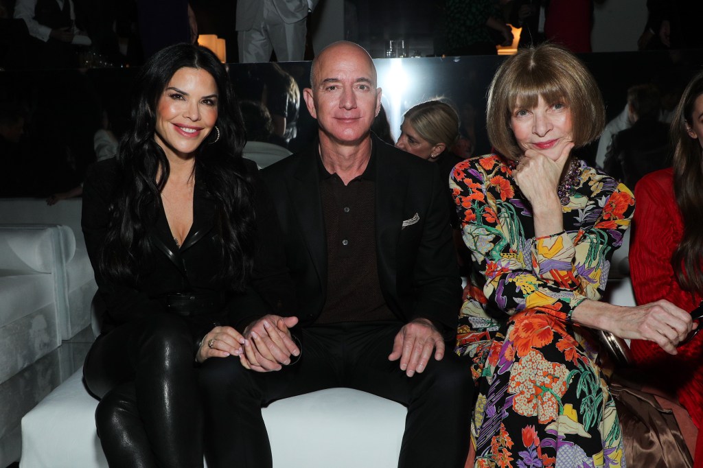 Lauren Sanchez, in a black top and black pants, Jeff Bezos in a black shirt and black pants, and Anna Wintour, in a floral dress, sitting in the front row at a fashion show.