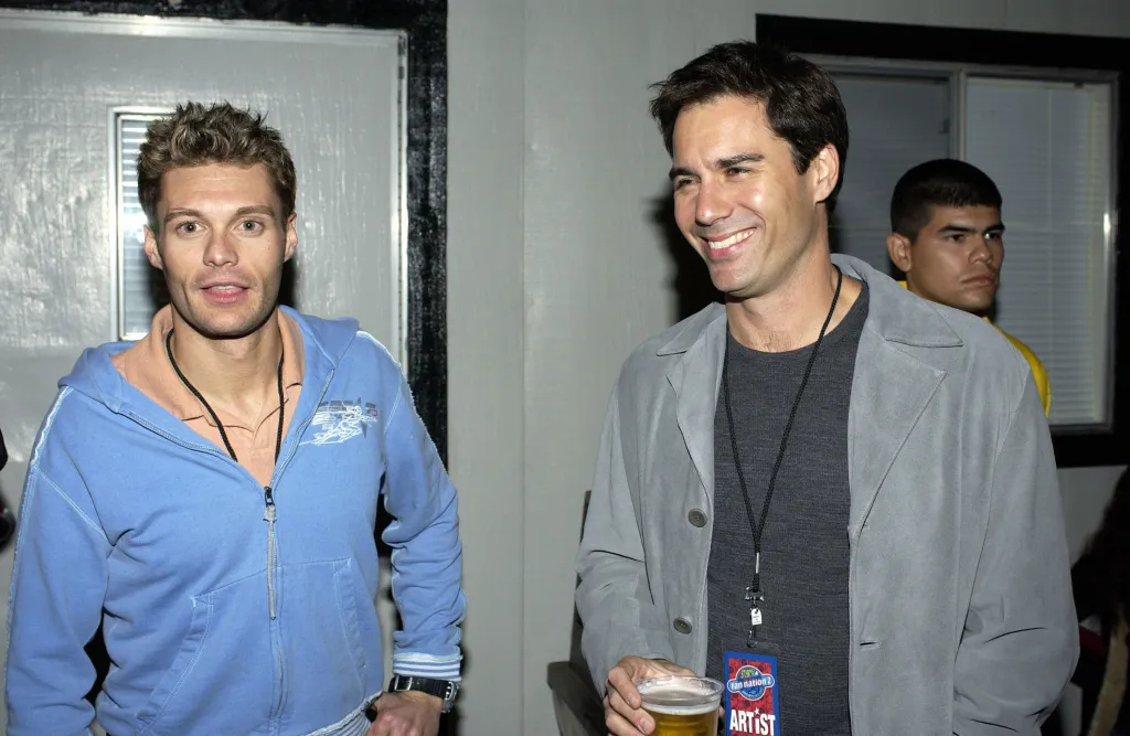 Ryan Seacrest in a blue hoodie and Eric McCormack in a gray jacket holding a drink, smiling.