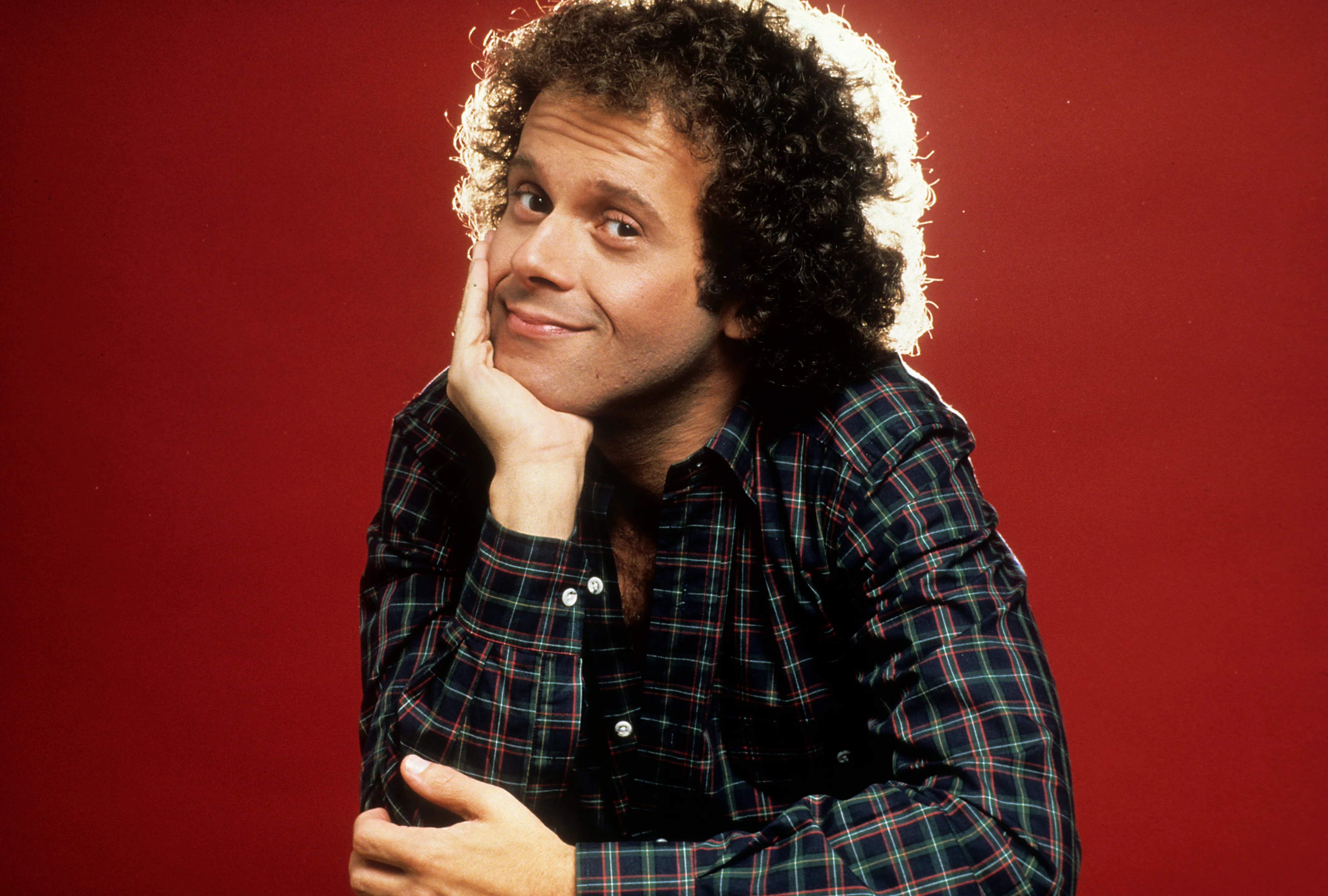 Richard Simmons in a plaid shirt with his hand on his chin.
