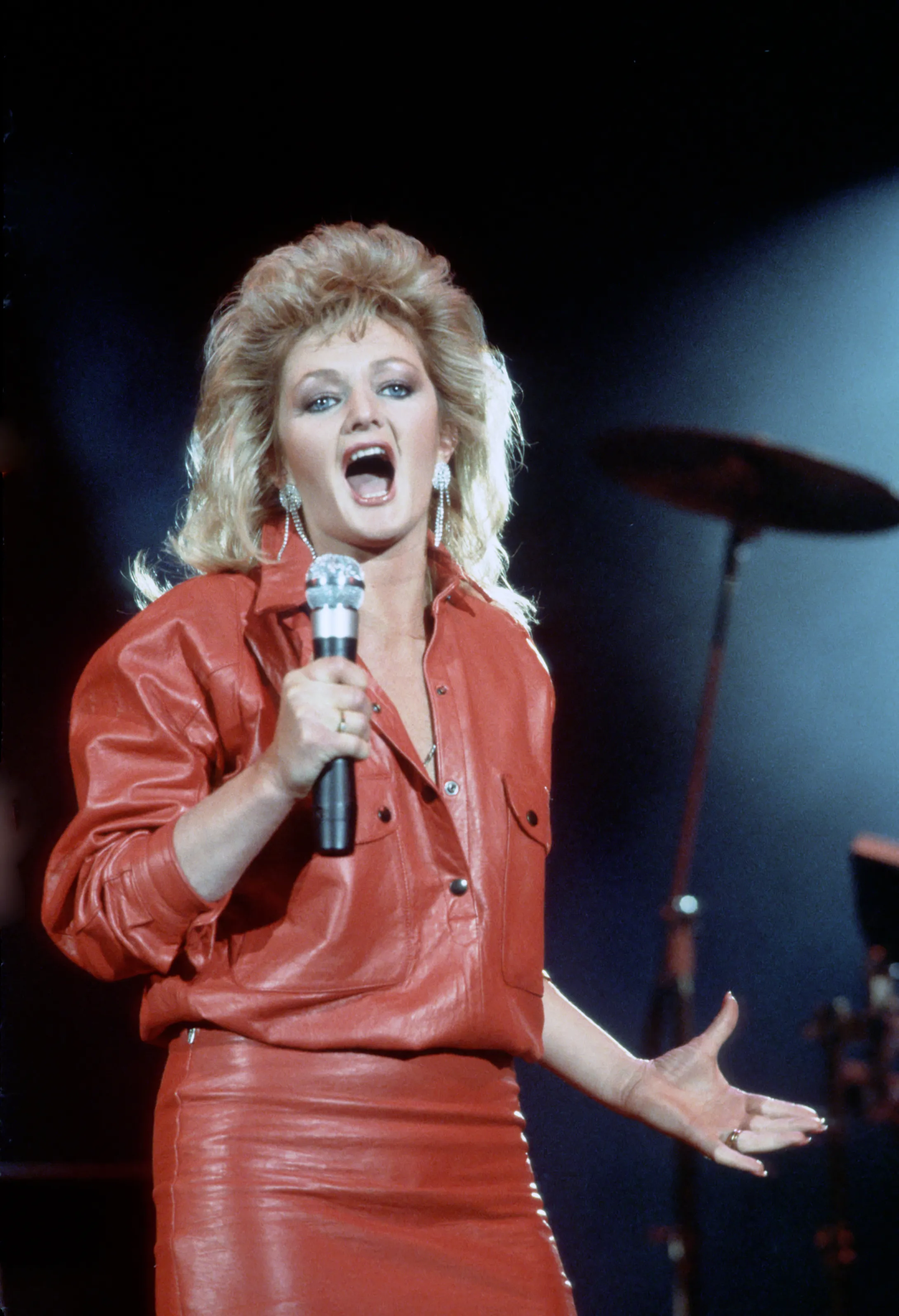 Bonnie Tyler singing into a microphone, wearing a red leather top and skirt.