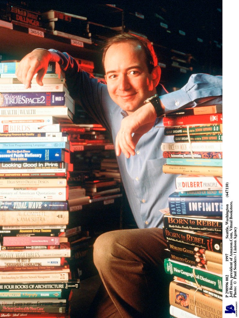 Jeff Bezos, founder of Amazon, leans on a stack of books with more books in the background. He wears a blue shirt and brown pants. 