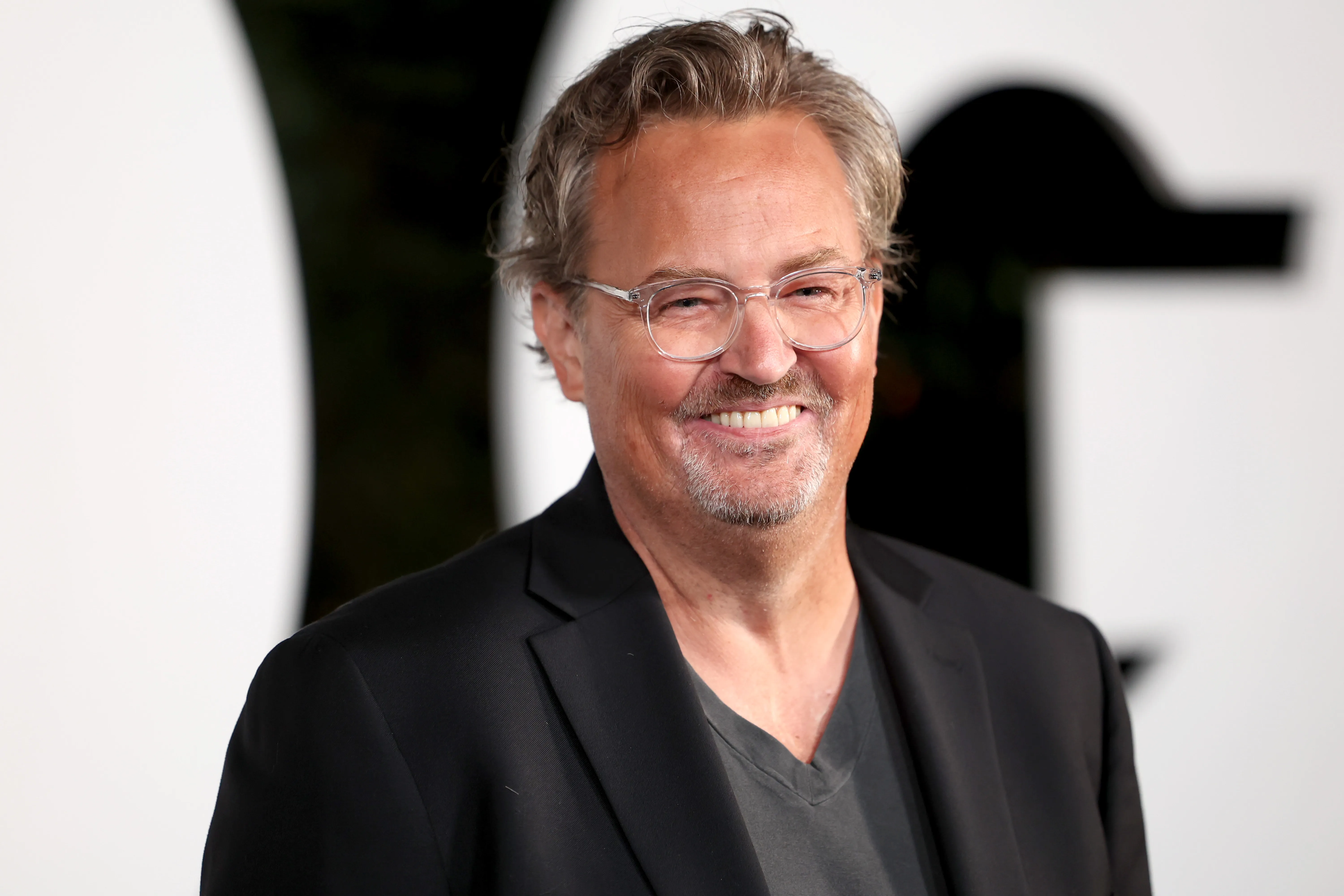 Actor Matthew Perry smiles at the 2022 GQ Men of the Year Party, wearing clear-rimmed glasses and a black blazer over a gray V-neck shirt.