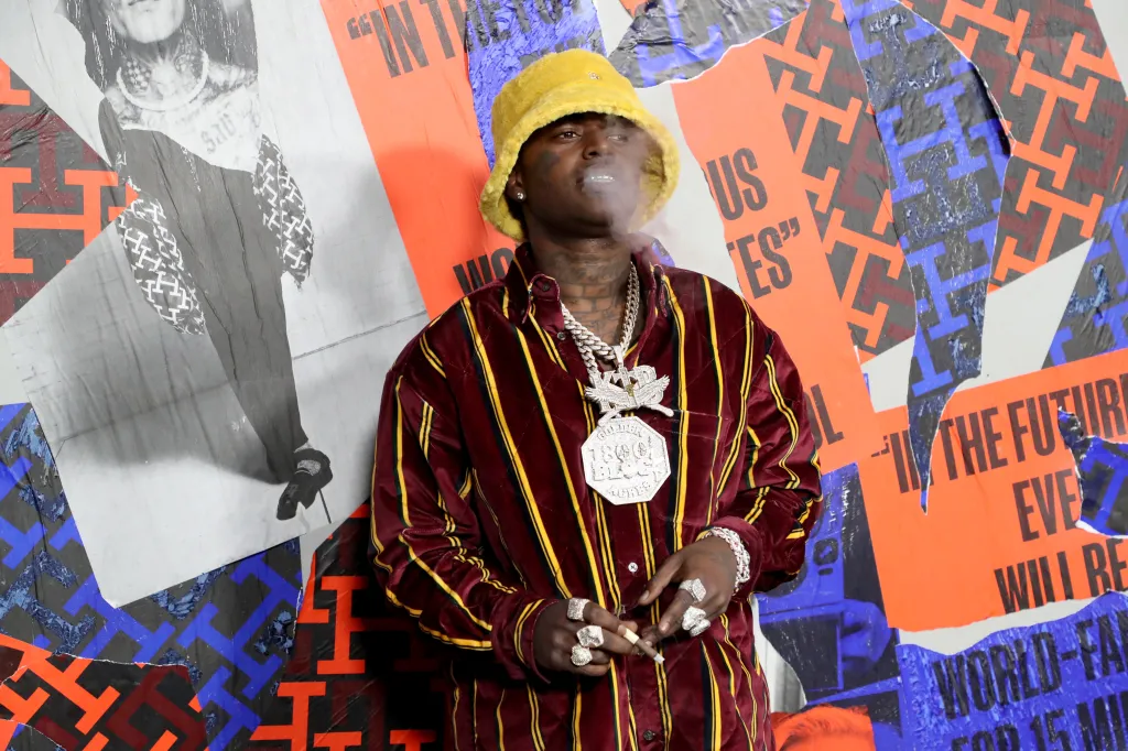 Kodak Black in a yellow fuzzy hat and a striped shirt, blowing smoke, at the Tommy Hilfiger fashion show.
