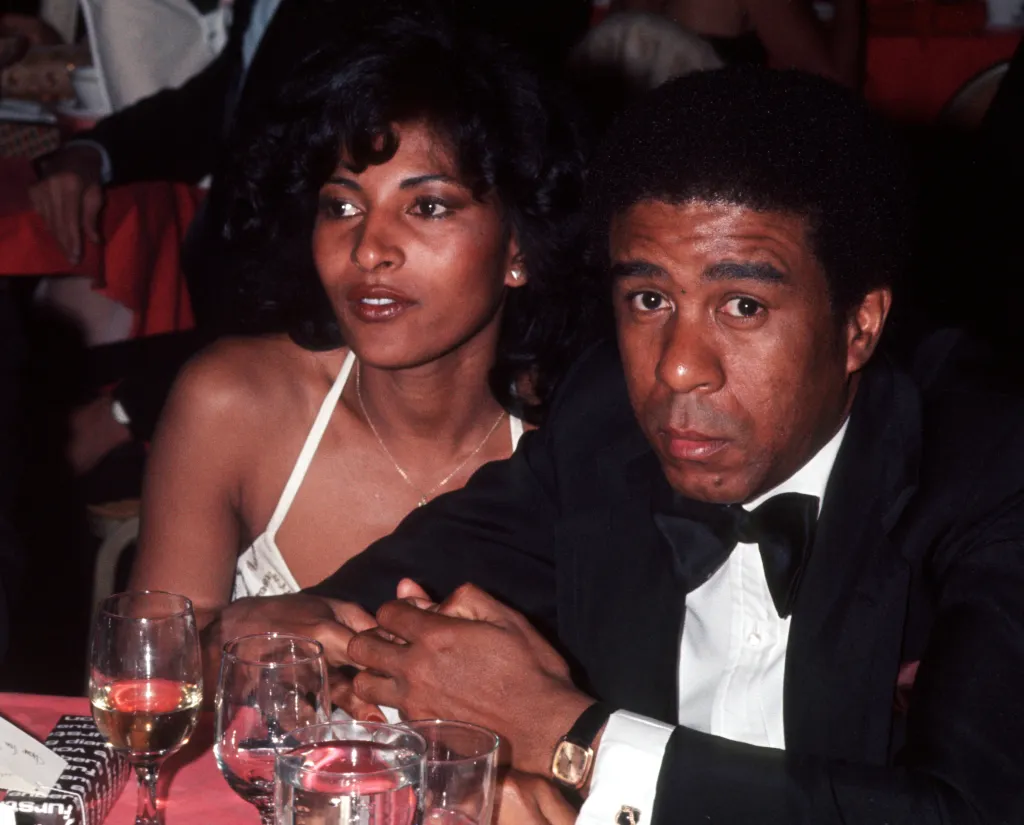 Pam Grier and Richard Pryor sit at a table, holding hands.