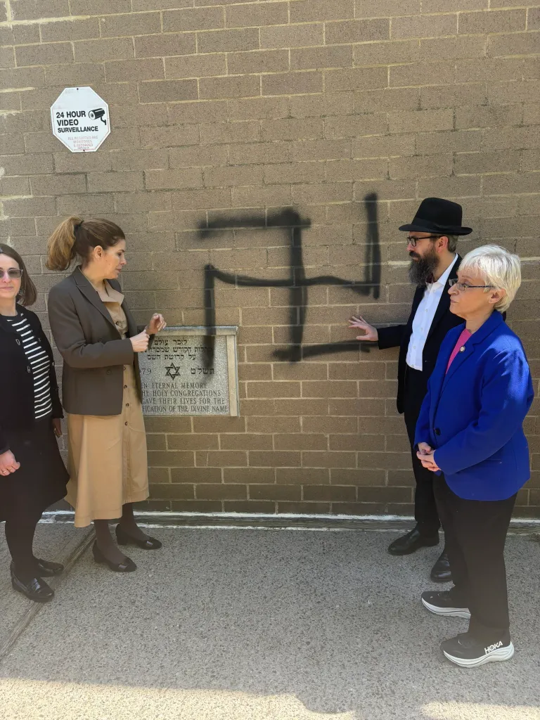 People standing next to antisemitic graffiti on the wall of Congregation Machane Chodosh.
