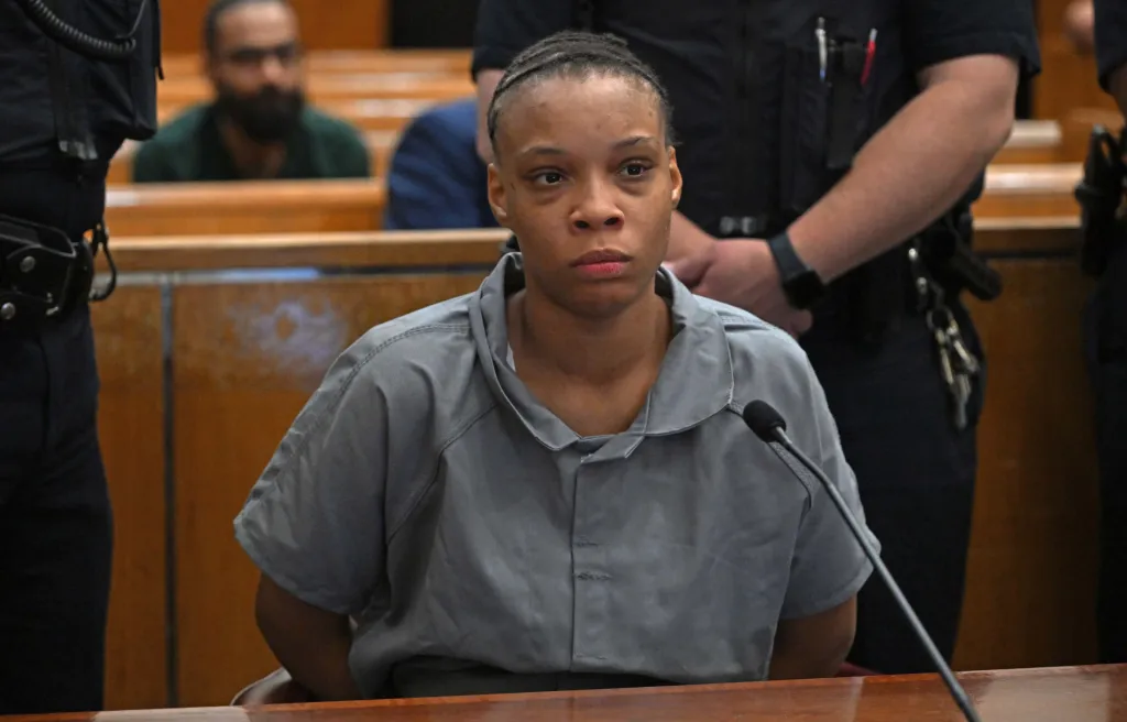 Naytavia Ragsdale in a grey shirt, seated at a wooden table in a courtroom, with a microphone in front of her.