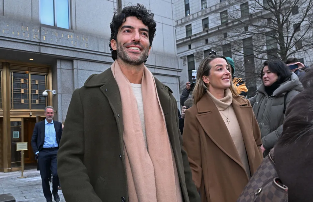 Justin Baldoni smiling, wearing a dark green coat and light pink scarf, leaving federal court with another woman.
