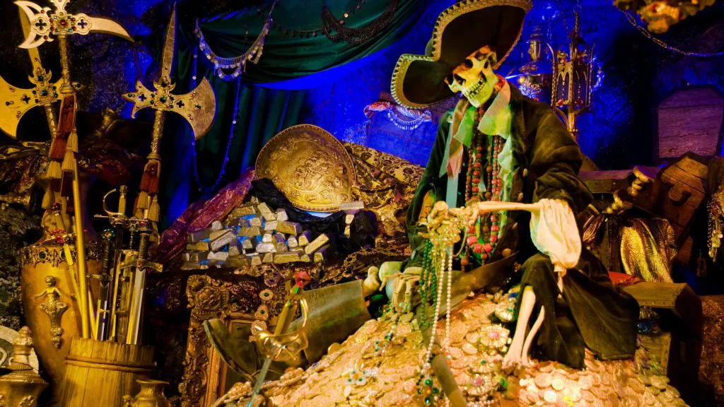 A skeleton wearing a pirate hat and coat sits atop a pile of gold, surrounded by treasure and weapons.