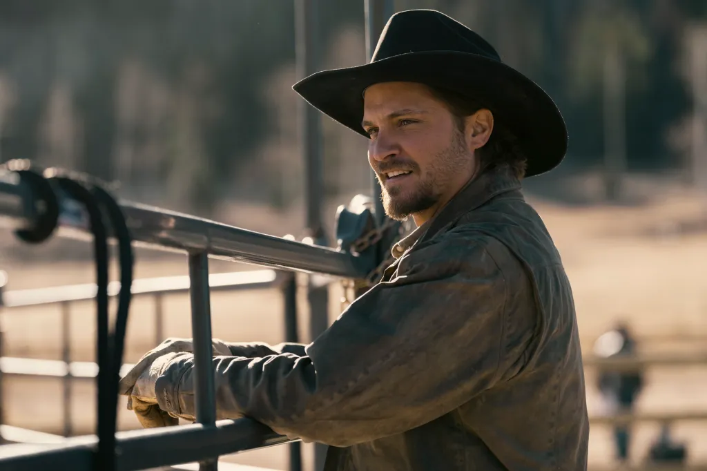 Luke Grimes as Kayce Dutton wearing a cowboy hat and gloves, leaning on a metal fence.