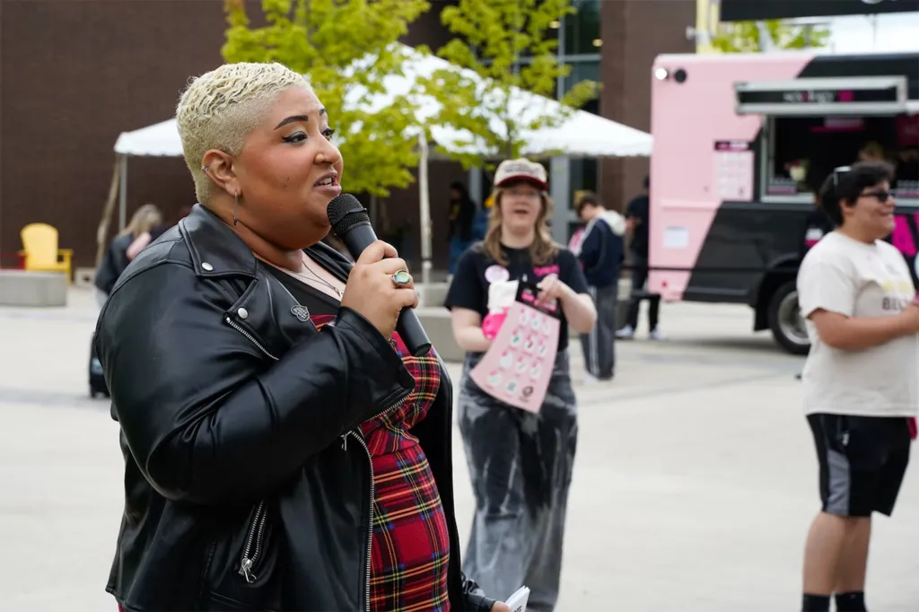 Dirty Lola, a pleasure expert, speaks into a microphone at an event with a pink food truck in the background.