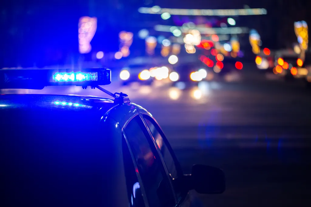 Blue police lights flashing on a police car at night with blurred city lights in the background.